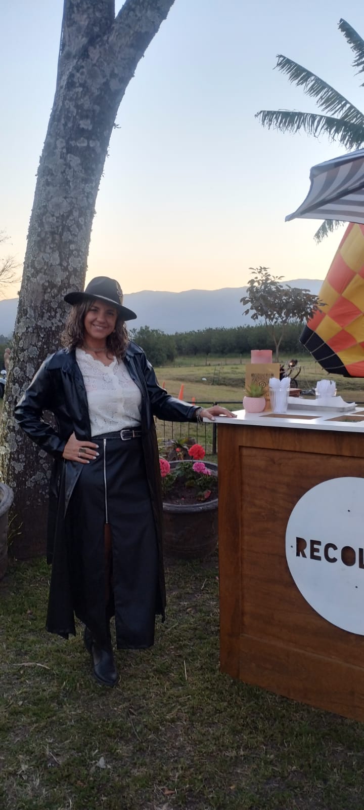 Mujer con sombrero de paño negro customizado por malala.tuc, saco largo de cuero y pollera negra, en un evento al aire libre al atardecer