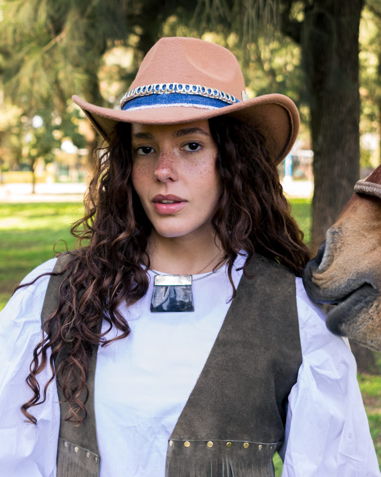 Modelo con sombrero de paño camel customizado con banda de jean y trenzado, junto a un caballo